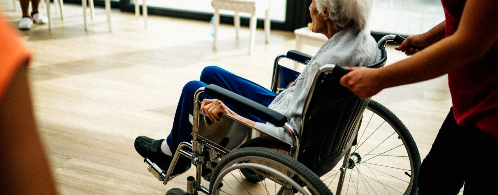 A caregiver pushes an older woman with white hair in a wheelchair across a bright day room with large windows, the kind of setting a PACE day center provides