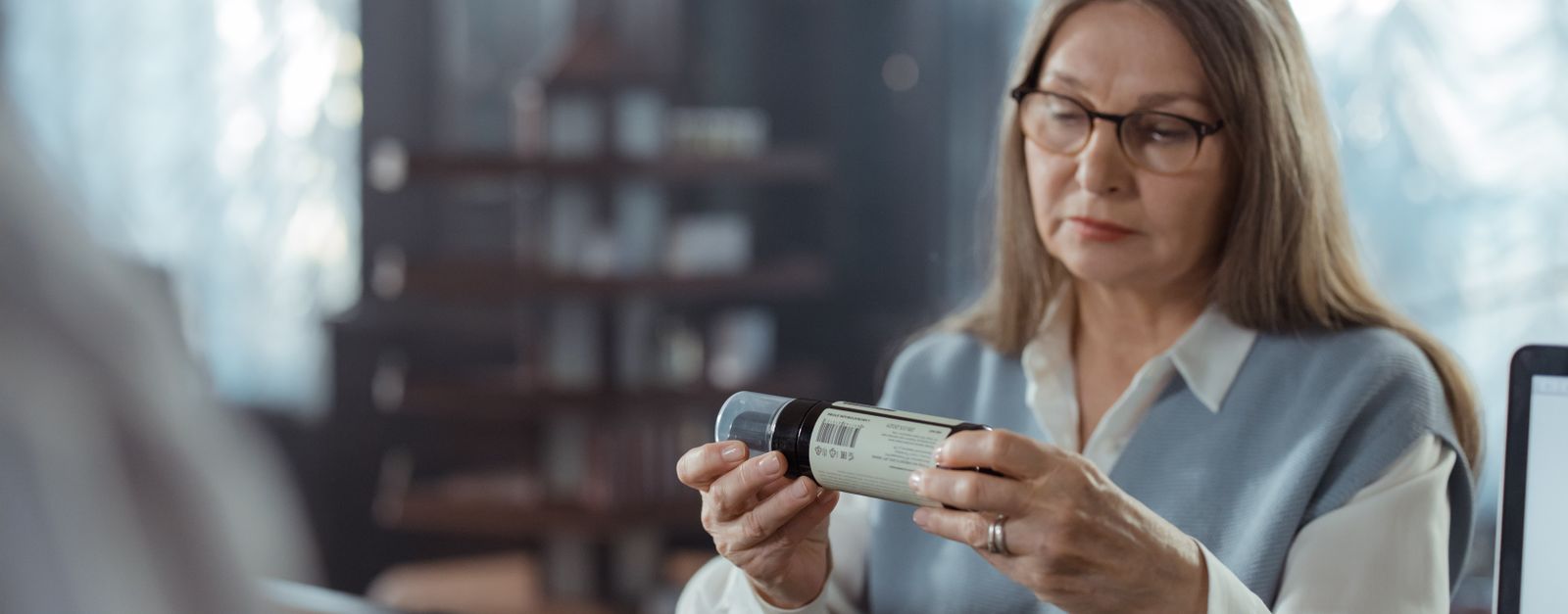 An older woman in glasses reading the label on a prescription bottle at a pharmacy counter while deciding whether to switch her Medicare Part D plan for 2027