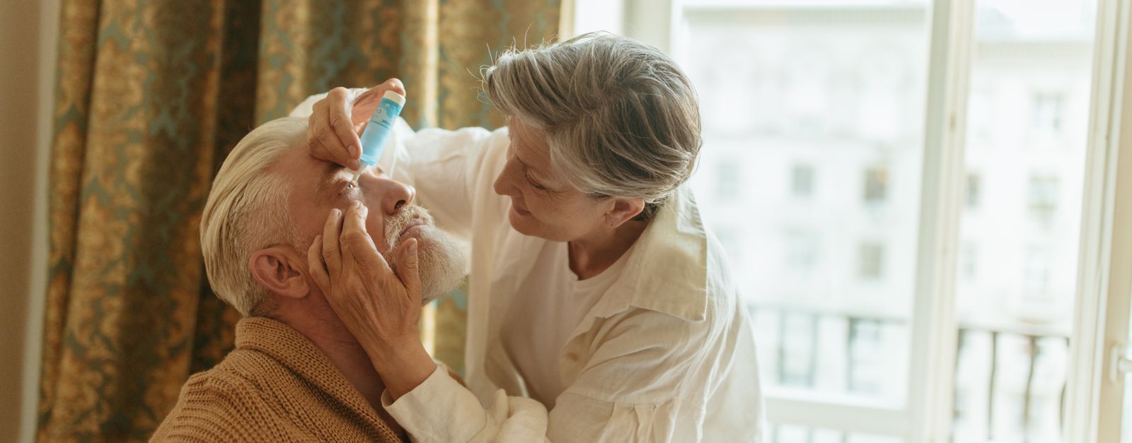 An older woman gently puts eye drops in her husband's eye at home, the kind of care that follows cataract surgery