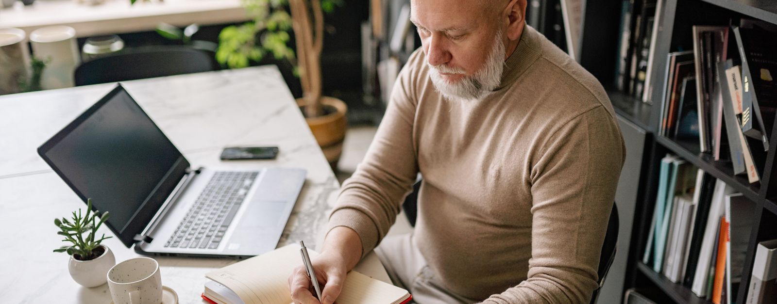 A gray-haired man writing notes in a notebook beside an open laptop, working through a comparison of Medicare Advantage plans for 2027