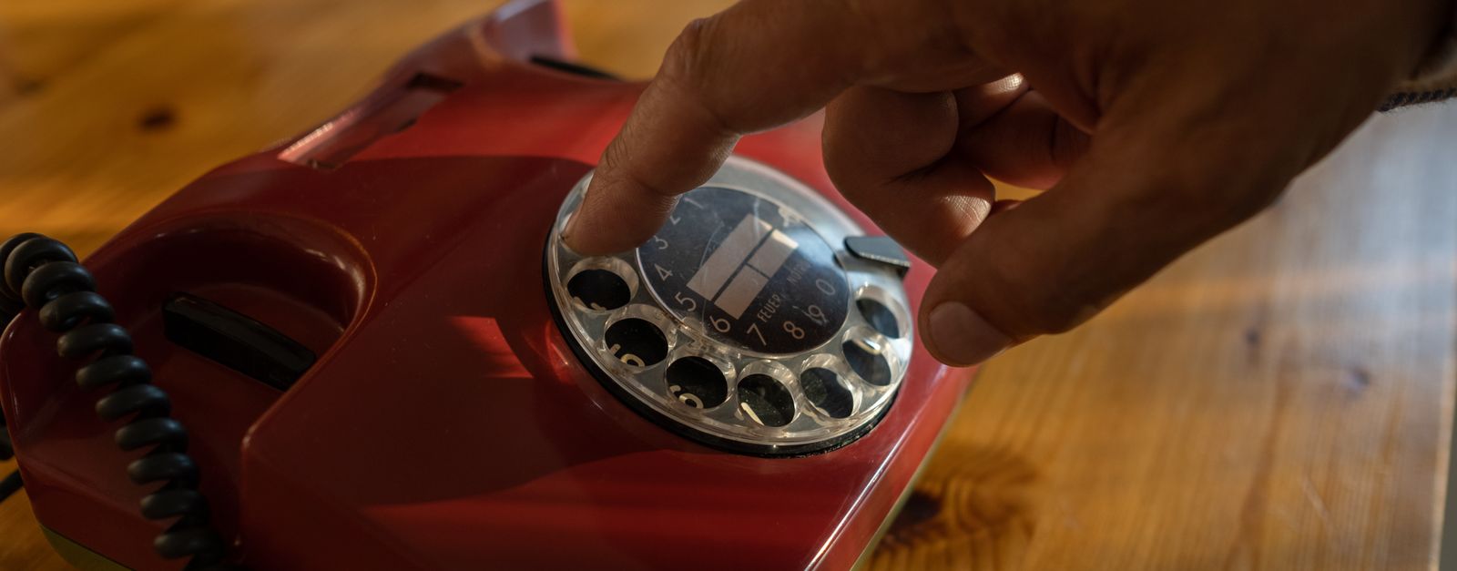 An older adult's hand dialing a red rotary telephone on a wooden table — calling back a number they trust instead of answering a stranger's call about a Presbyterian Medicare plan