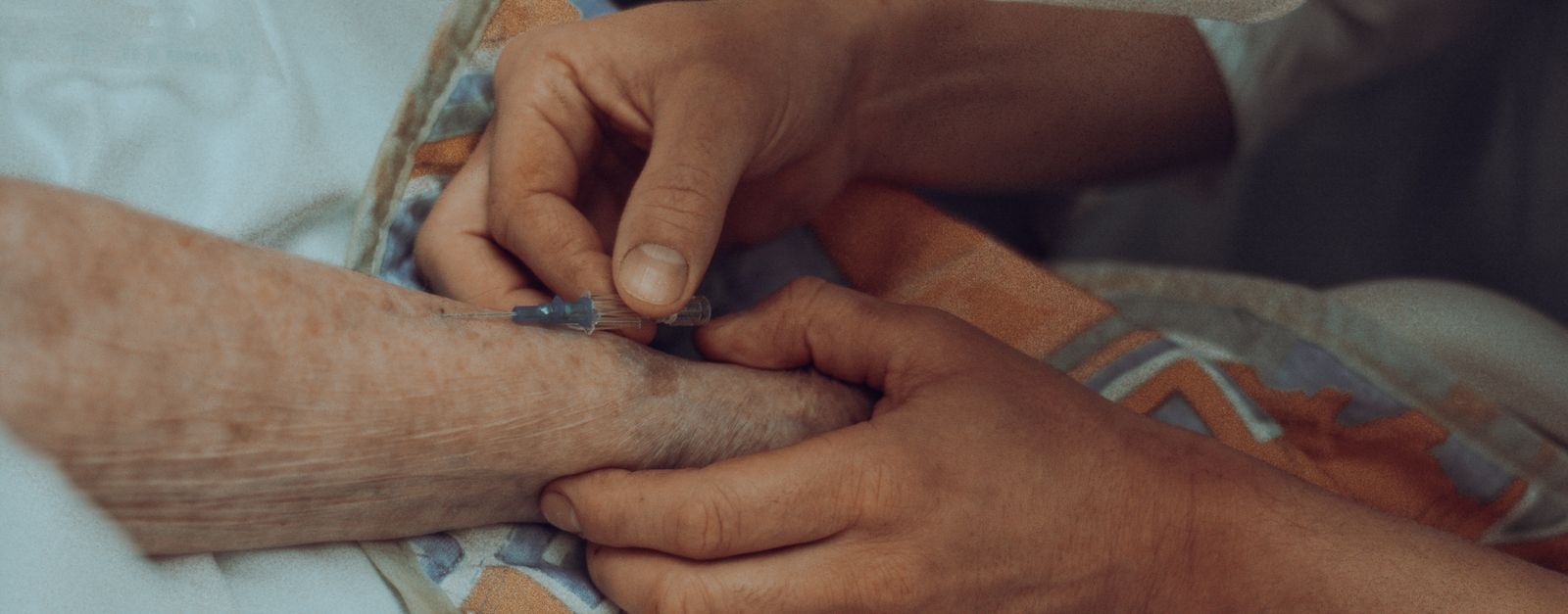 A caregiver's hands holding an older patient's hand with an IV line in place — a New Mexico Medicare member in the middle of treatment as the Presbyterian Medicare Advantage plan ends