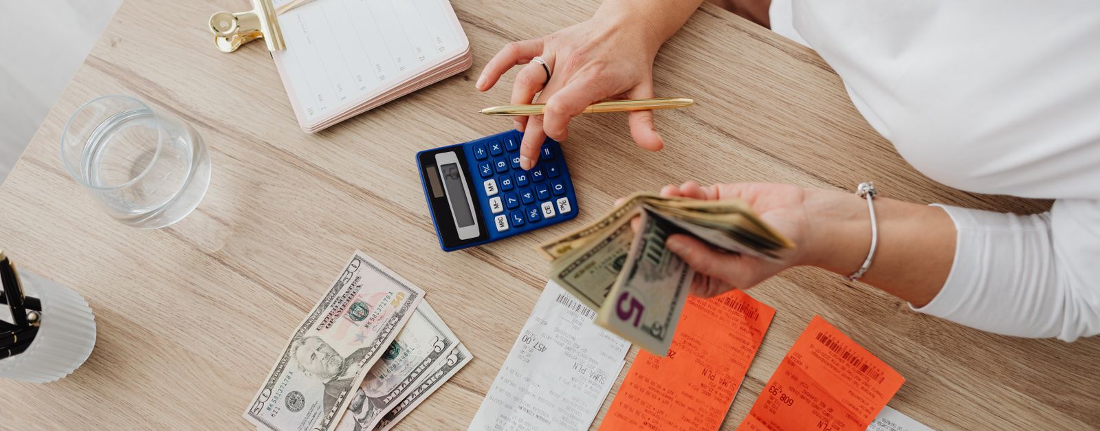 Seen from above, a woman's hands work a calculator at a wooden table with receipts, a notebook and a few bills spread out — adding up what a $0 premium Medicare Advantage plan costs over a year